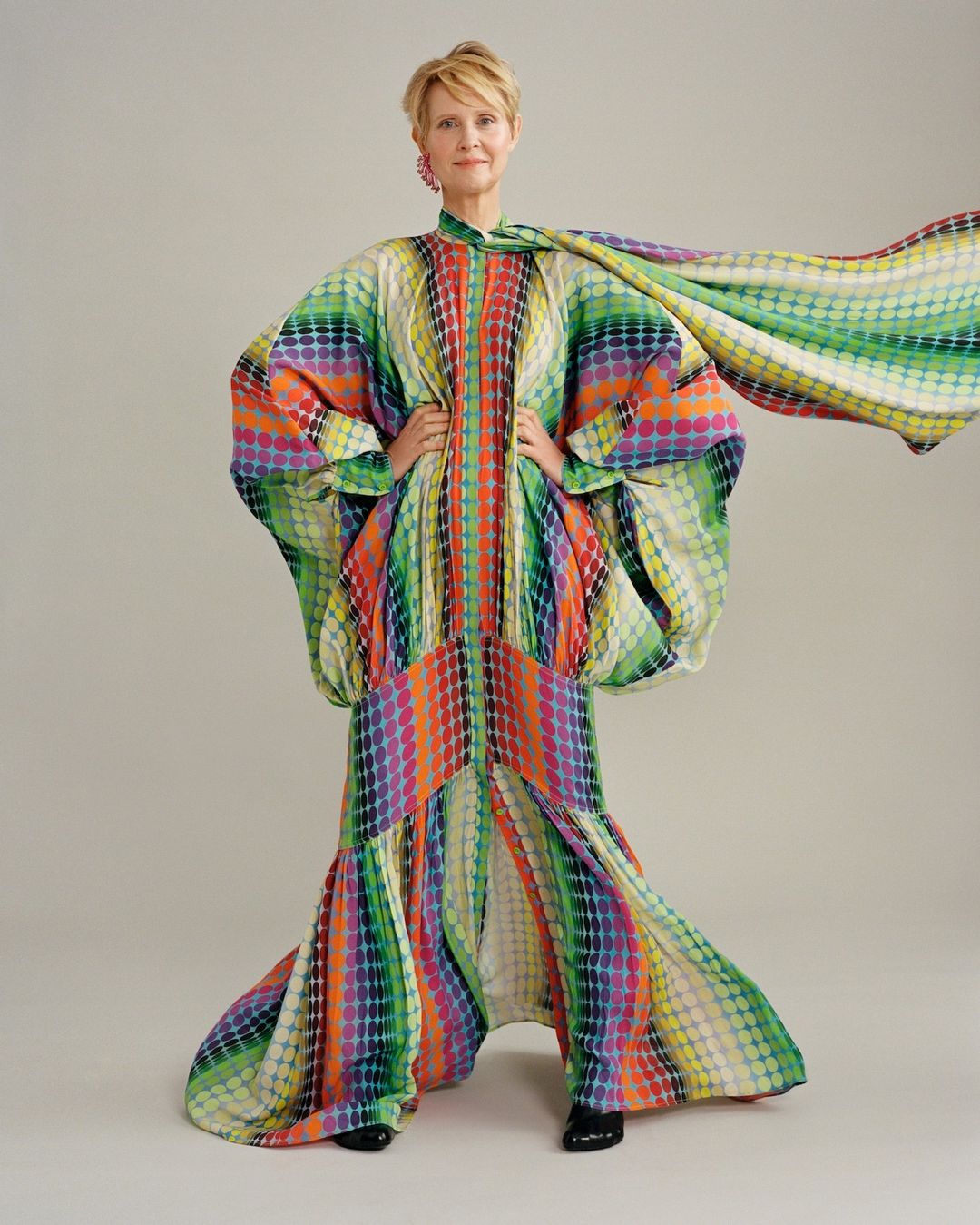 Adult woman in flowing gown with colorful dot pattern poses in a studio, looking at the camera with hands on her waist.