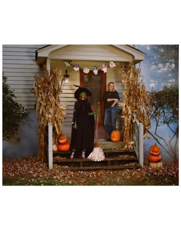 A woman in witch costume stands on a porch with Halloween pumpkins, while another adult stands on the house's porch stairs.