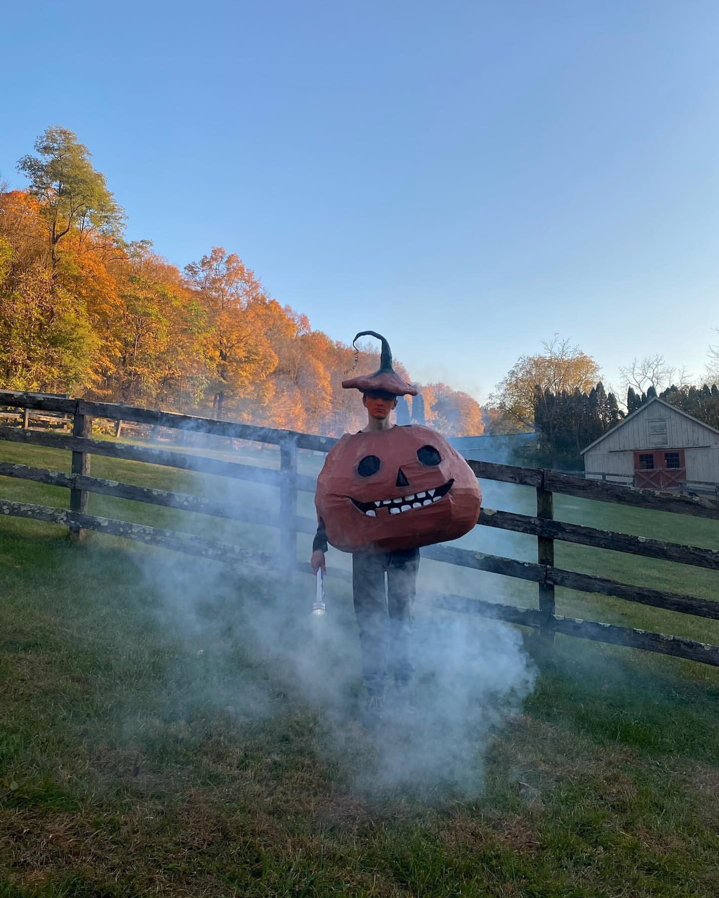 Person in a pumpkin costume stands in a grassy field with smoke, a wooden fence, autumn trees, and a barn in the background.