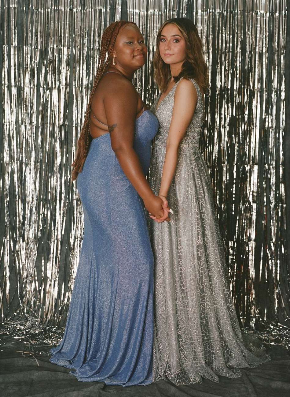Two women in formal dresses hold hands in front of a metallic backdrop. Prom or special occasion fashion portrait.
