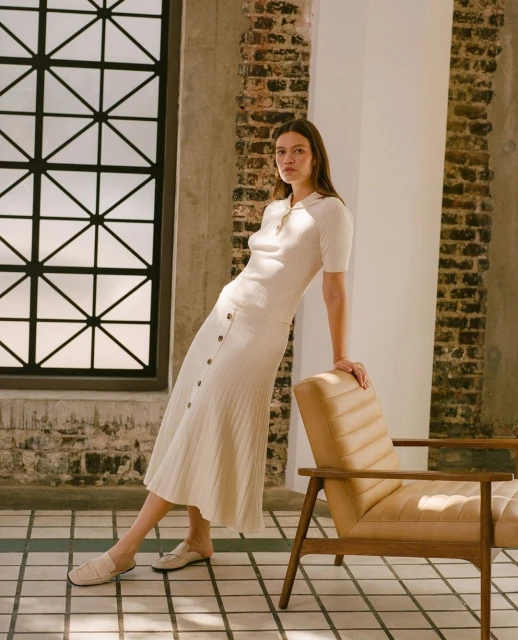 Brunette woman in a knit top and skirt leans against a wall with a window, posing next to a tan mid-century modern armchair.