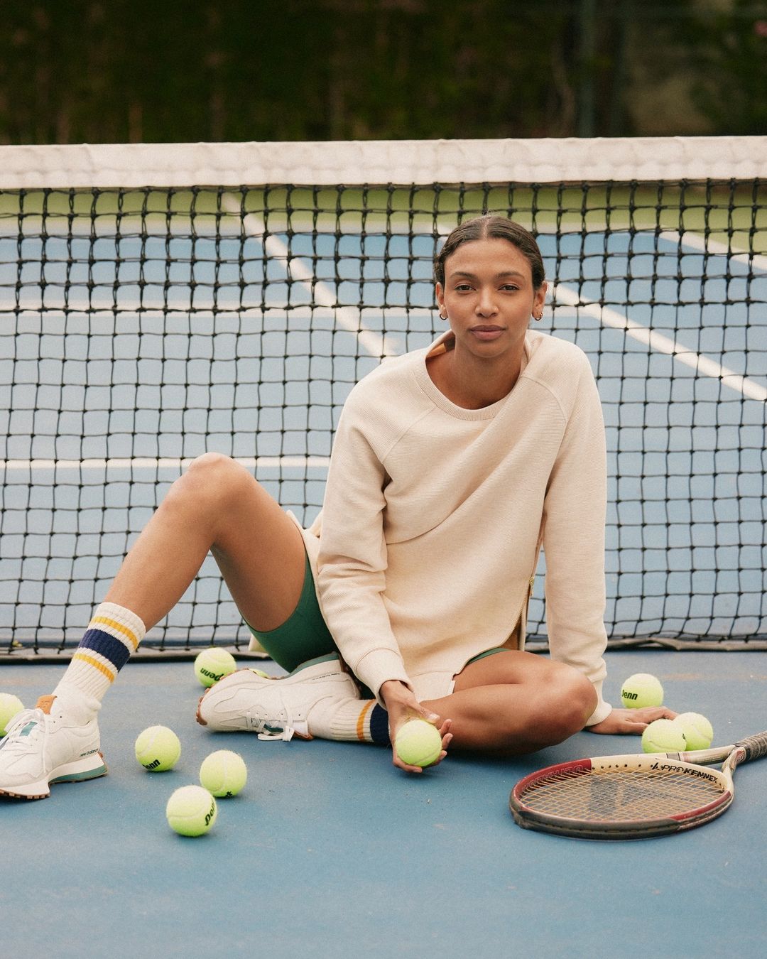 Woman sitting on a blue tennis court with balls and a racket wearing a sweatshirt and shorts with a net and trees in the background.