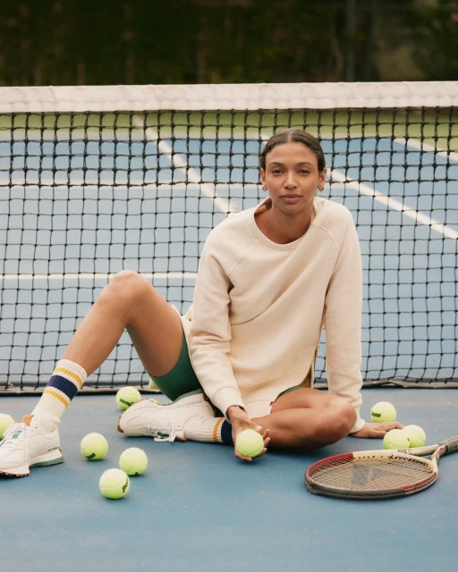 Woman sitting on a blue tennis court with balls and a racket wearing a sweatshirt and shorts with a net and trees in the background.