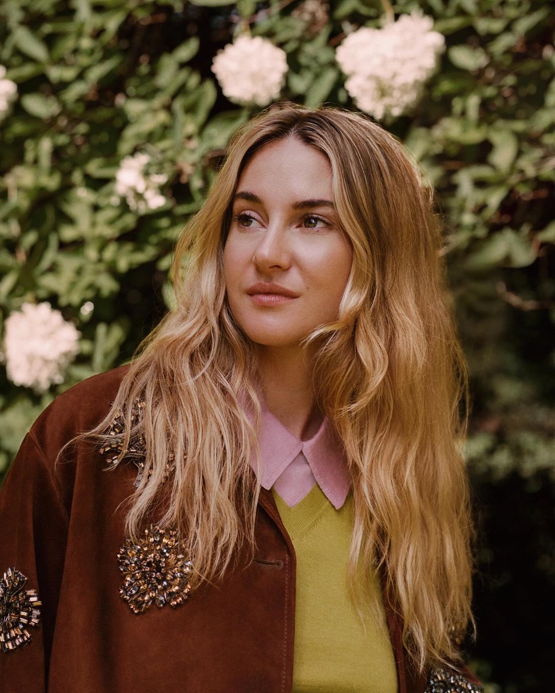 Blonde woman in a brown jacket with beaded embellishments poses in front of a leafy green background with white flowers in an outdoor setting.