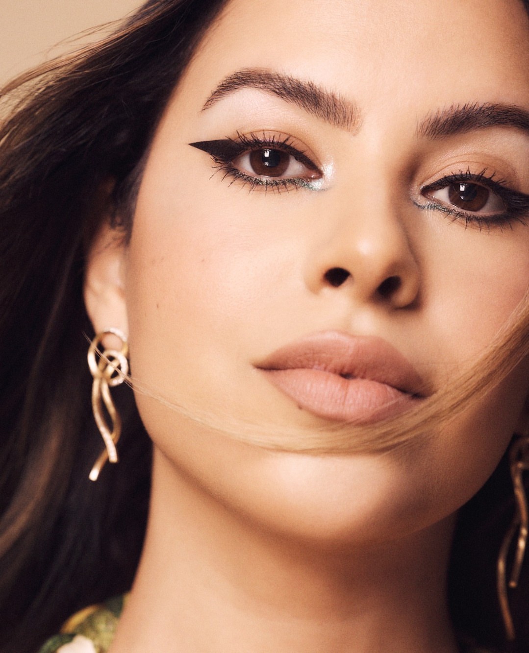 Close-up portrait of a young woman with brown eyes, heavy black eyeliner, and dangling gold earrings against a neutral background.