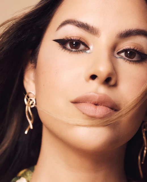 Close-up portrait of a young woman with brown eyes, heavy black eyeliner, and dangling gold earrings against a neutral background.