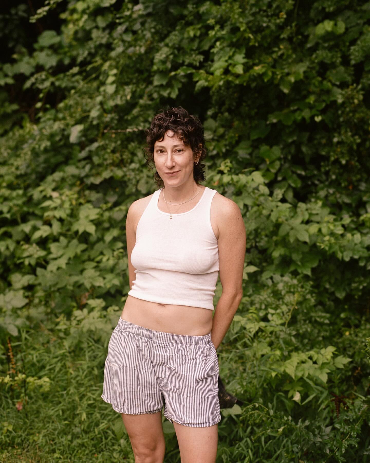 Portrait of a woman wearing a white tank top and striped shorts standing outdoors against a dense green foliage background.