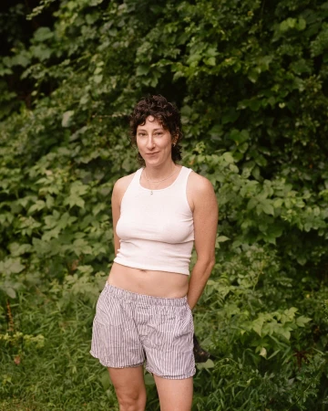 Portrait of a woman wearing a white tank top and striped shorts standing outdoors against a dense green foliage background.