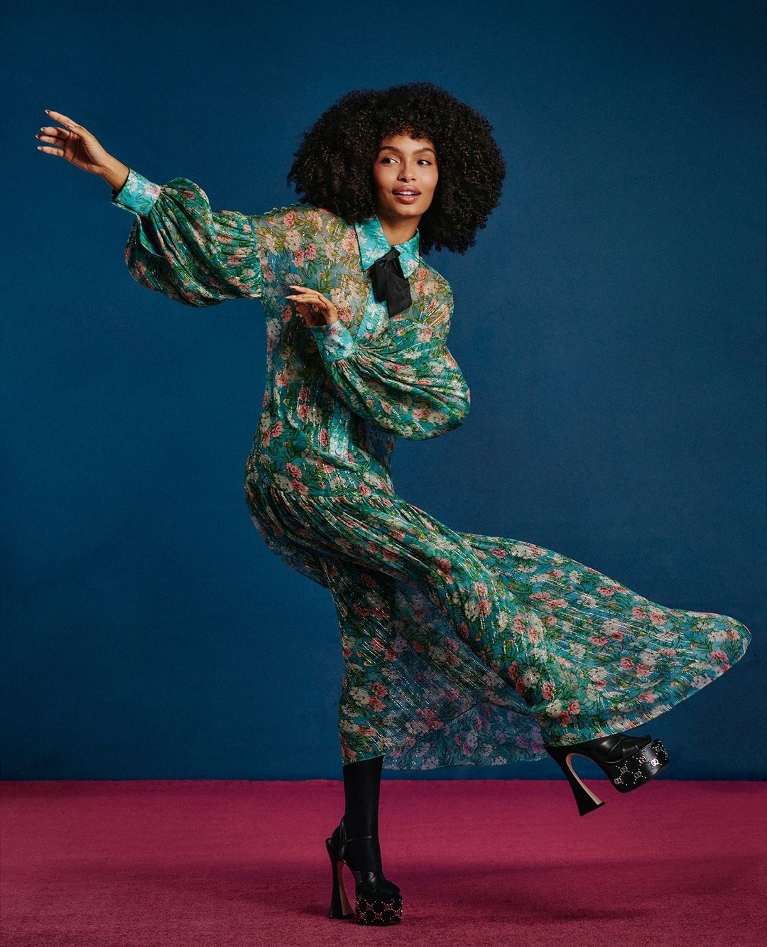 A woman with afro-style hair, wearing a green floral dress and high-heeled platform shoes, poses in a studio with a blue backdrop.