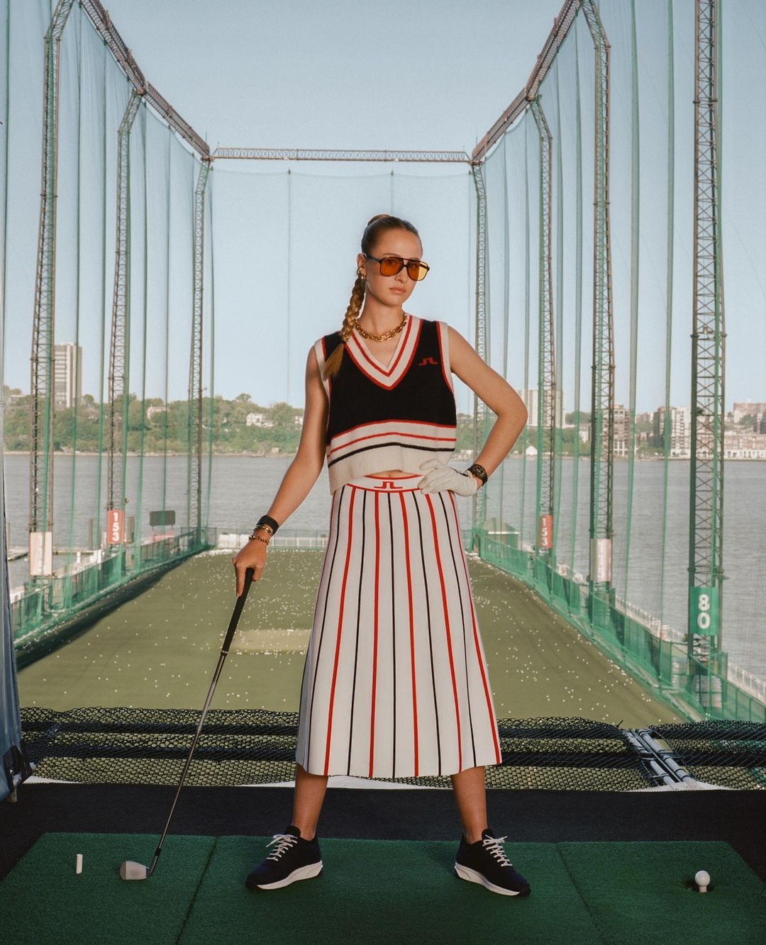 A woman in a golf skirt and sweater vest poses with a golf club on a driving range against a backdrop of a cityscape and water.