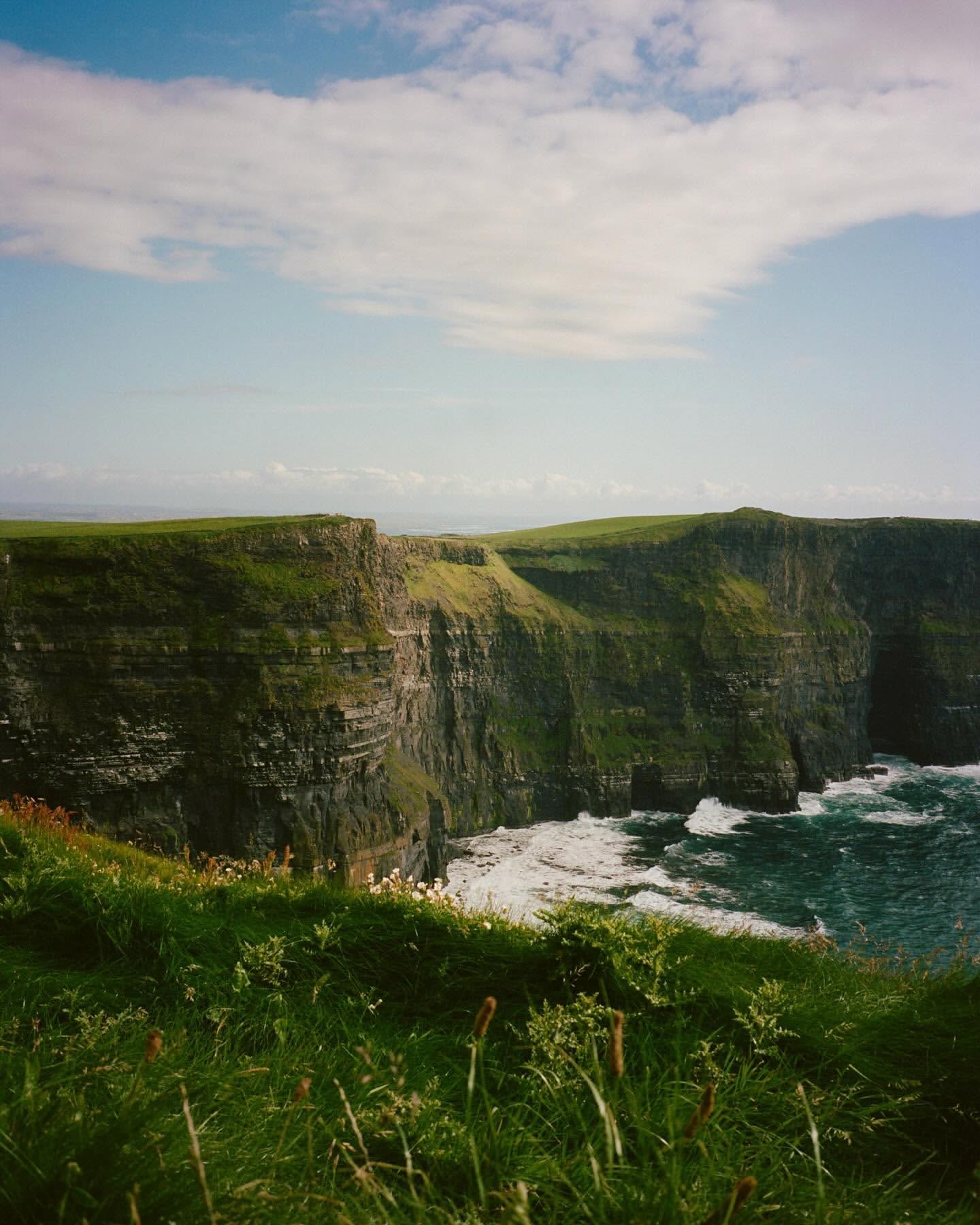 Cliffs of Moher coastline view featuring green grass, rock cliffs, crashing waves, and a blue sky with scattered clouds in Ireland.