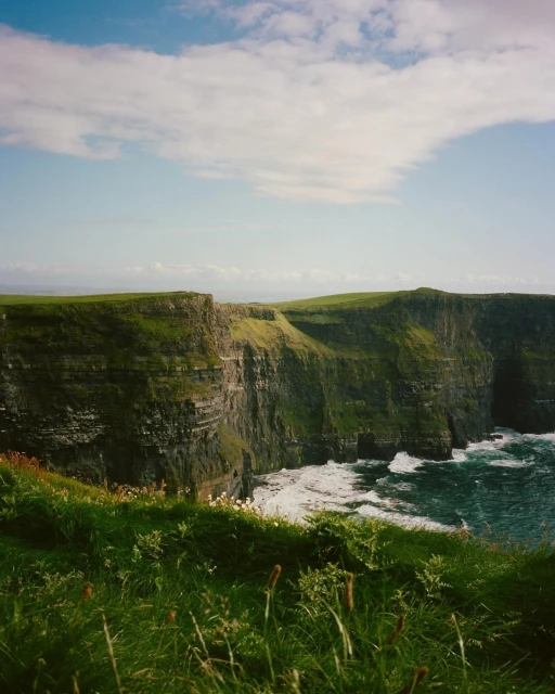 Cliffs of Moher coastline view featuring green grass, rock cliffs, crashing waves, and a blue sky with scattered clouds in Ireland.