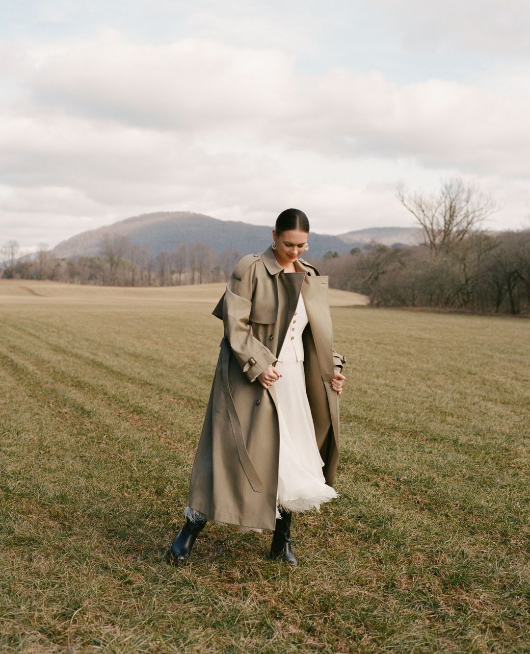 A woman wearing a long trench coat and skirt walks across a dry winter field with rolling hills and bare trees in the background.