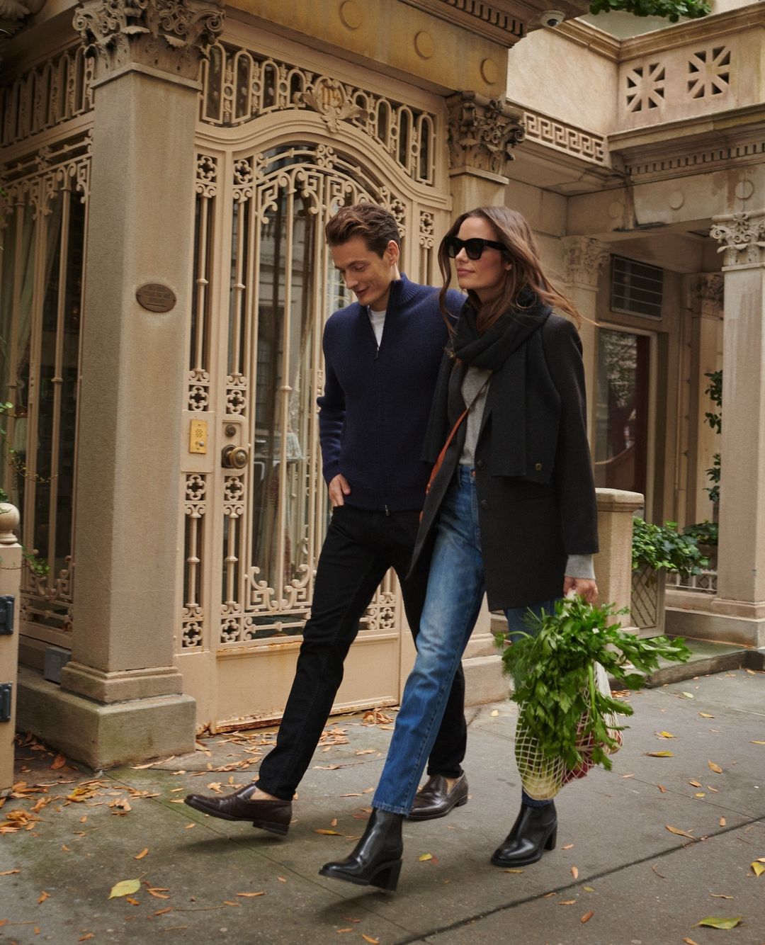 A stylish couple walking on a city street, with the woman carrying a bag of vegetables and both wearing fashionable outerwear.