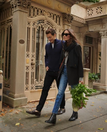 A stylish couple walking on a city street, with the woman carrying a bag of vegetables and both wearing fashionable outerwear.