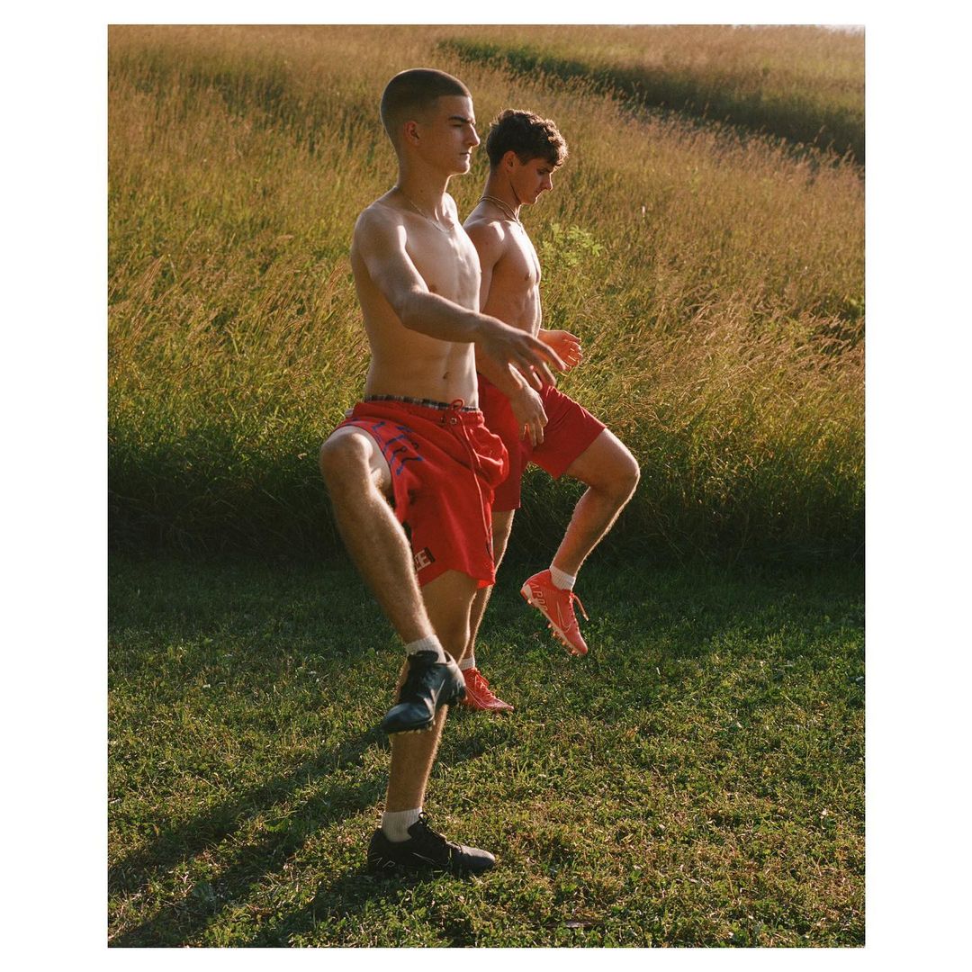 Two shirtless male athletes in red shorts perform dynamic leg stretches on a grassy field in the sunlight.
