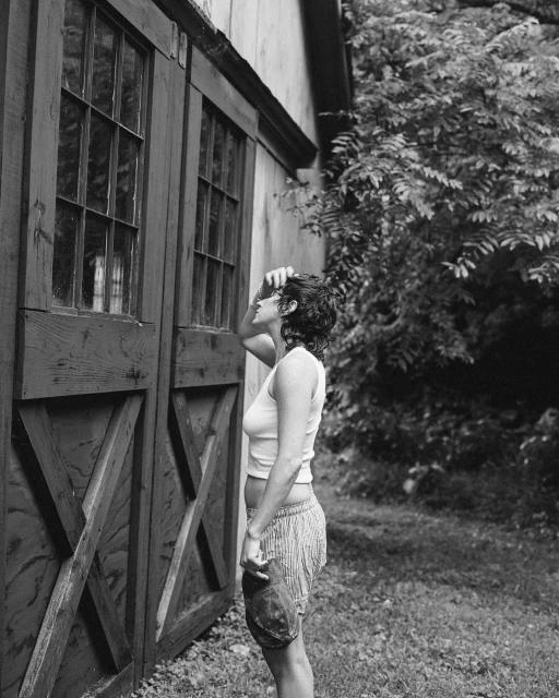 Black and white outdoor shot of a woman standing by a barn-style building with trees in the background, gazing upwards and shielding her eyes.