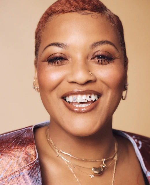 Close-up portrait of a smiling adult woman with short textured hair wearing layered necklaces, a metallic jacket, and dental jewelry.