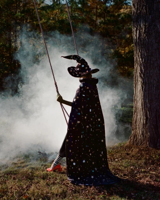 Person in witch costume on a rope swing in a forest with hazy smoke, wearing black cape and hat with star pattern, Halloween.