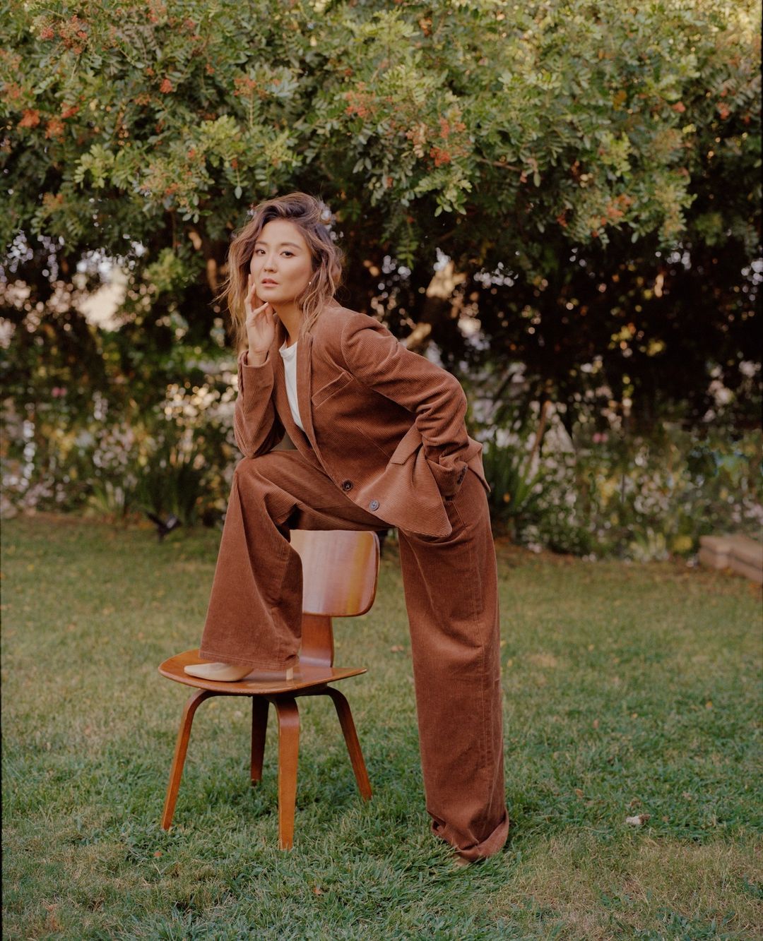 An adult woman poses on a wooden chair in a garden, wearing a brown corduroy suit, exuding a relaxed and casual elegance in an outdoor setting.