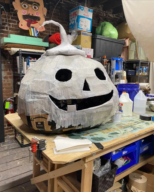 A large, handmade paper-mache jack-o'-lantern rests on a wooden table in a cluttered indoor workshop with shelves and brick walls.
