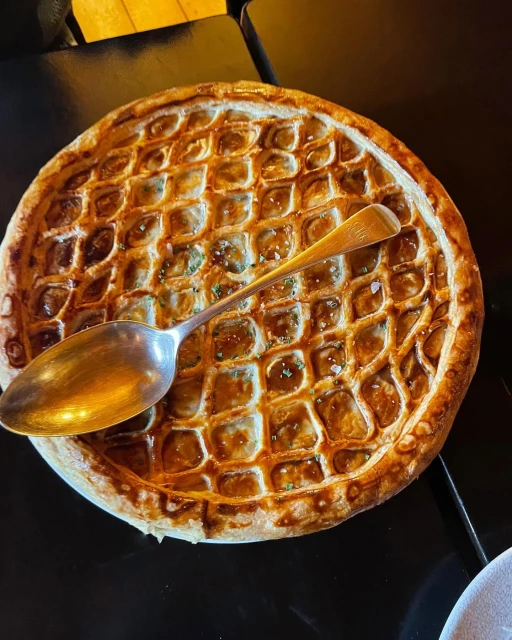 Overhead shot of a round pie with golden lattice crust, topped with a silver spoon on a dark surface. Food photography.