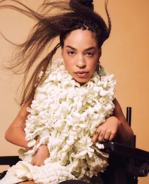 Portrait of a stylish adult female with textured hair and elaborate white ruffled collar, seated in wheelchair, against tan backdrop.