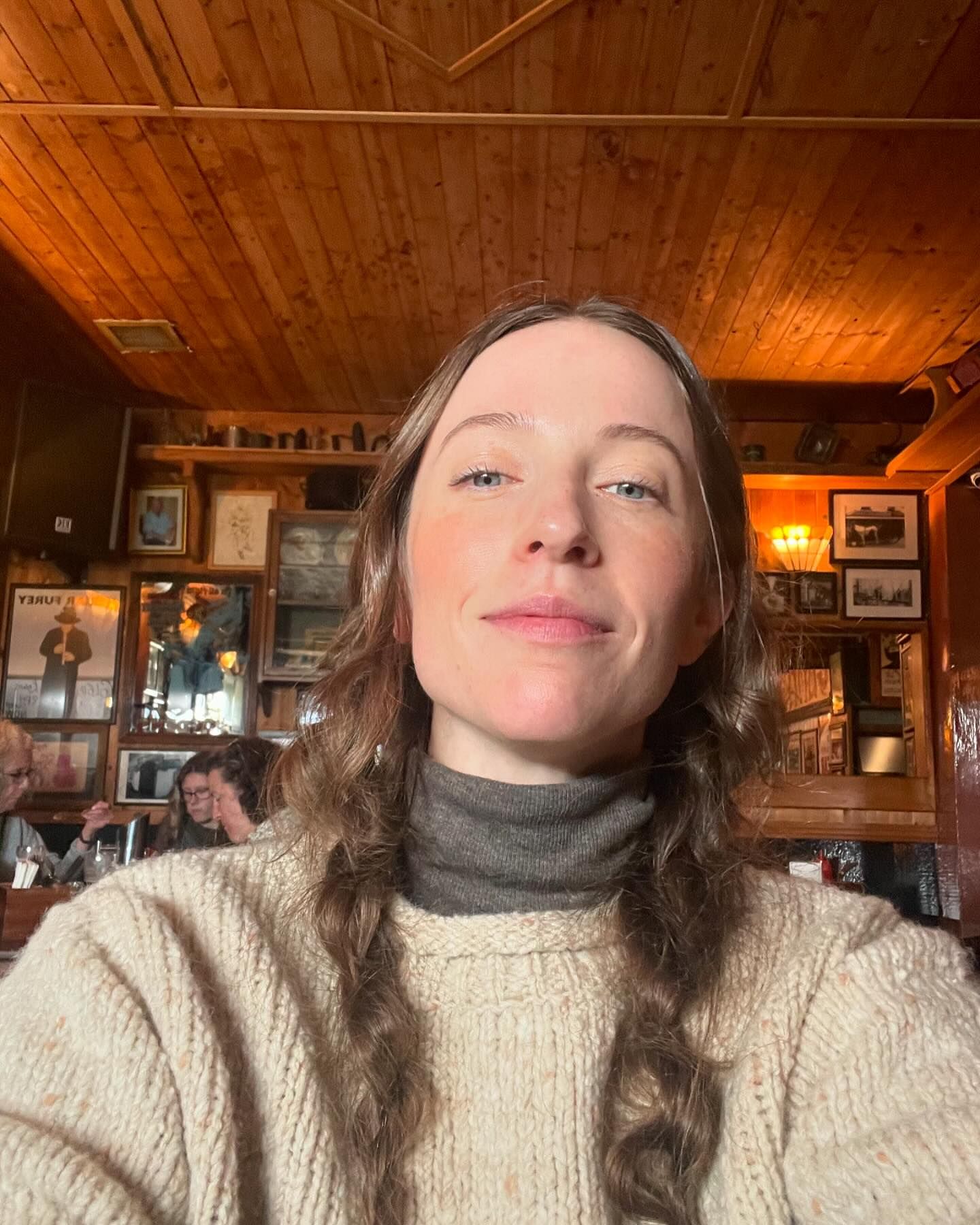 Woman in a cream-colored sweater takes a selfie indoors, with a wooden-paneled wall decorated with framed pictures in the background.