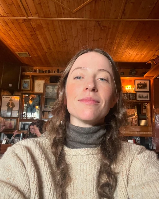 Woman in a cream-colored sweater takes a selfie indoors, with a wooden-paneled wall decorated with framed pictures in the background.
