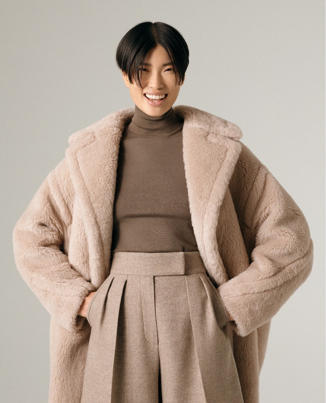 Smiling Asian woman posing in studio with a teddy-bear style coat, a brown turtleneck sweater, and wide-leg trousers in a fashion shot.