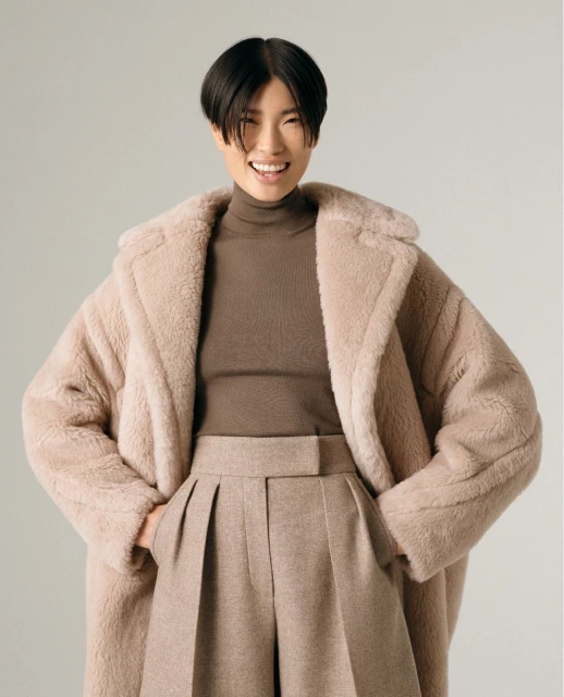 Smiling Asian woman posing in studio with a teddy-bear style coat, a brown turtleneck sweater, and wide-leg trousers in a fashion shot.