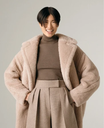 Smiling Asian woman posing in studio with a teddy-bear style coat, a brown turtleneck sweater, and wide-leg trousers in a fashion shot.