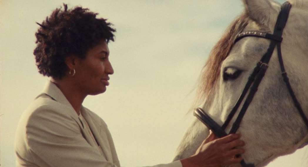 African American woman in profile, wearing a suit, touching the head of a white horse outdoors with a sky background in natural light.