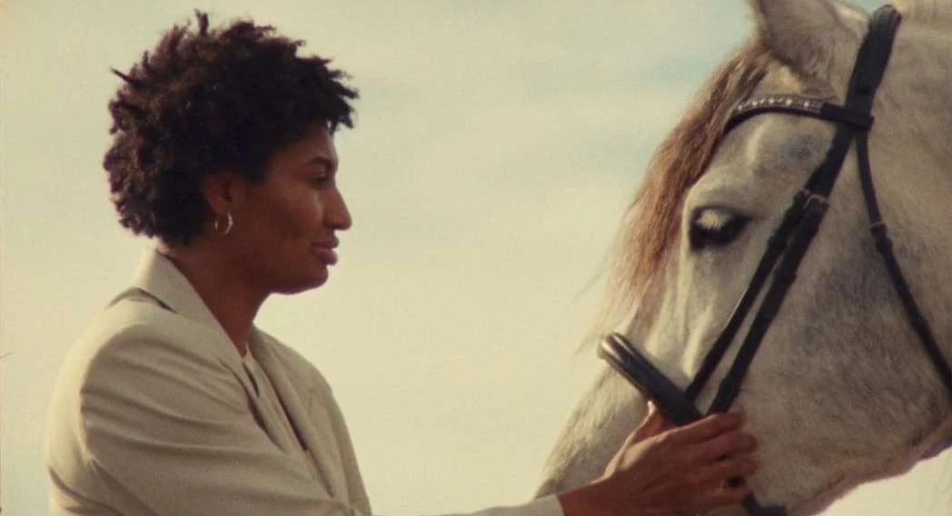 African American woman in profile, wearing a suit, touching the head of a white horse outdoors with a sky background in natural light.