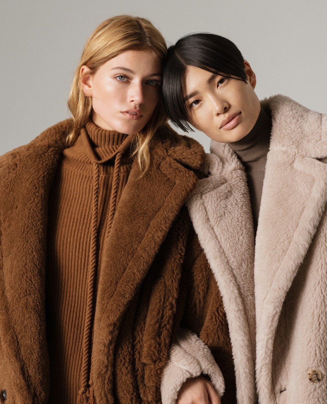 Two women in fashionable coats pose indoors, showcasing textures and neutral tones in a close-up shot for a fashion editorial.