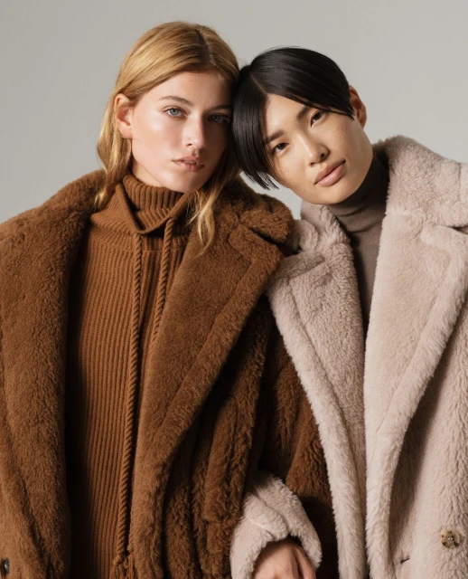 Two women in fashionable coats pose indoors, showcasing textures and neutral tones in a close-up shot for a fashion editorial.