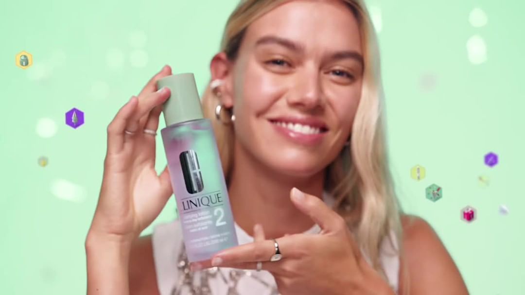 Smiling blonde woman presenting Clinique clarifying lotion in a studio with mint background, featuring brand logo and pastel-colored shapes.