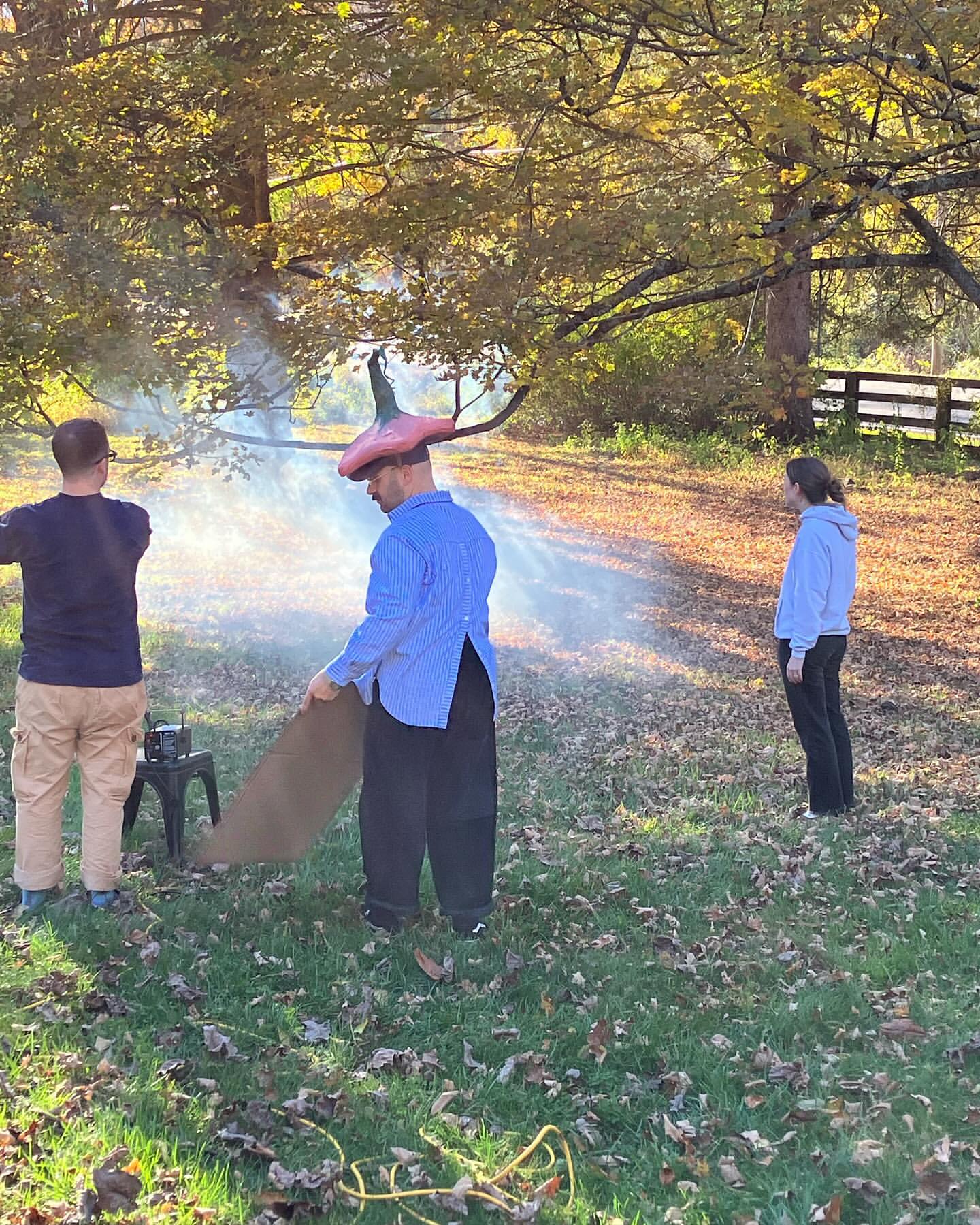 Outdoor scene with three people and a smoke machine, creating a hazy atmosphere in a grassy area with trees and a wooden fence.