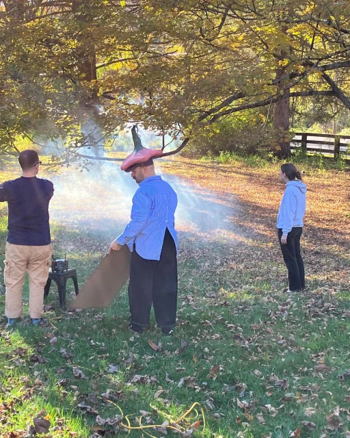 Outdoor scene with three people and a smoke machine, creating a hazy atmosphere in a grassy area with trees and a wooden fence.