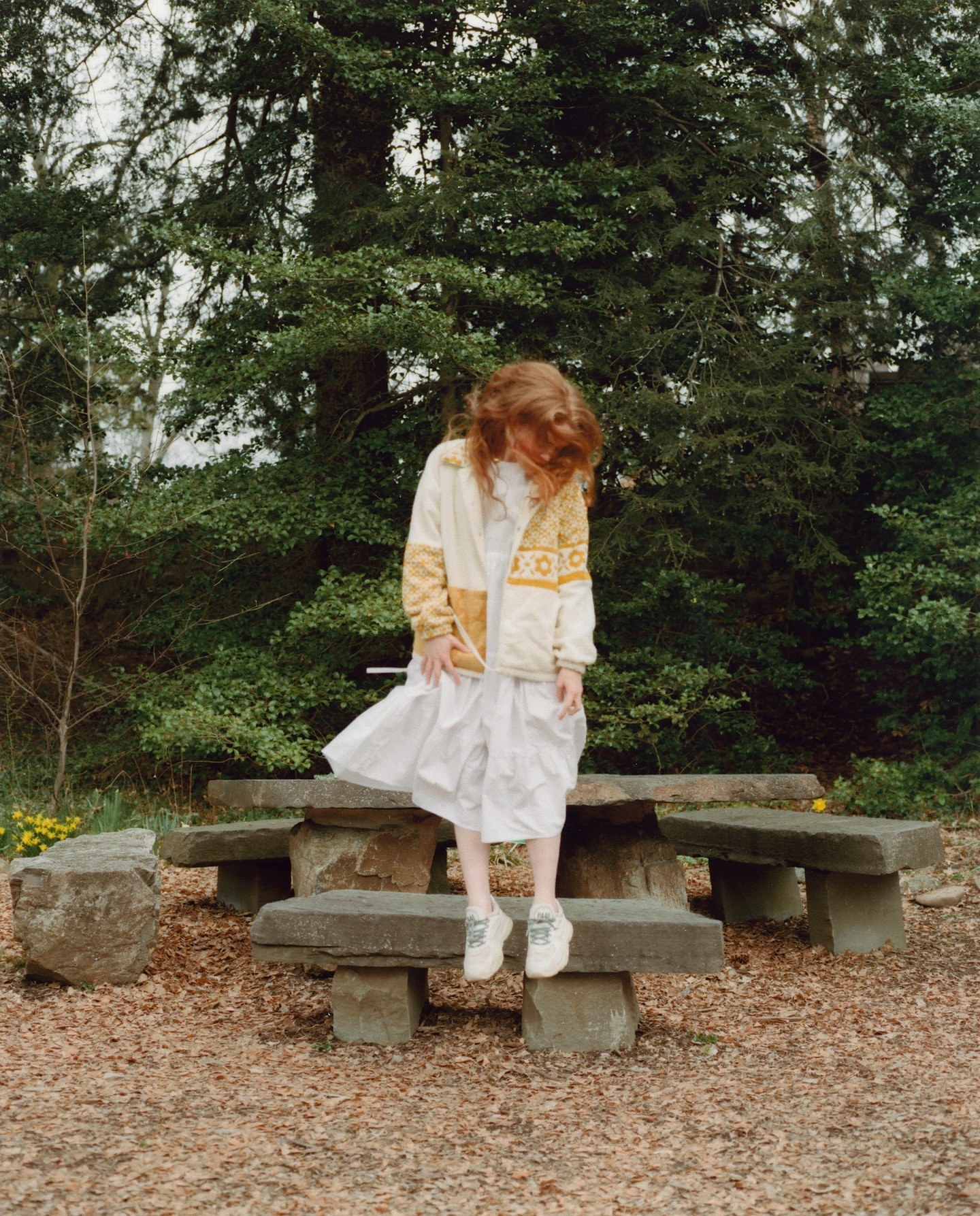 Child in white dress sits on stone table outdoors. Red-haired girl wears jacket near leafy green trees in a park.