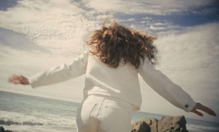 Rear view of a person with long curly hair wearing a white jacket and white pants, arms outstretched outdoors by the ocean.