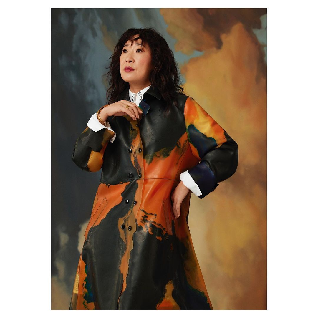 Sandra Oh poses in front of a painted backdrop, wearing a colorful patterned coat and white collared shirt with a neutral expression.