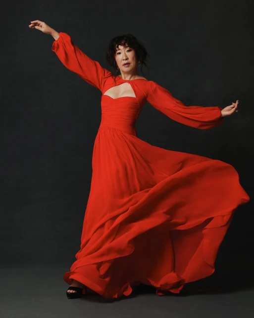 Woman in a red flowing gown, posing with outstretched arms in a photo studio. Full body shot against a neutral background, beauty and fashion photography.