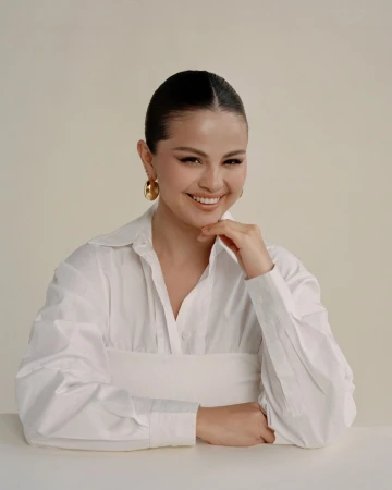 Selena Gomez wearing a white button-down shirt and gold hoop earrings against a neutral background
