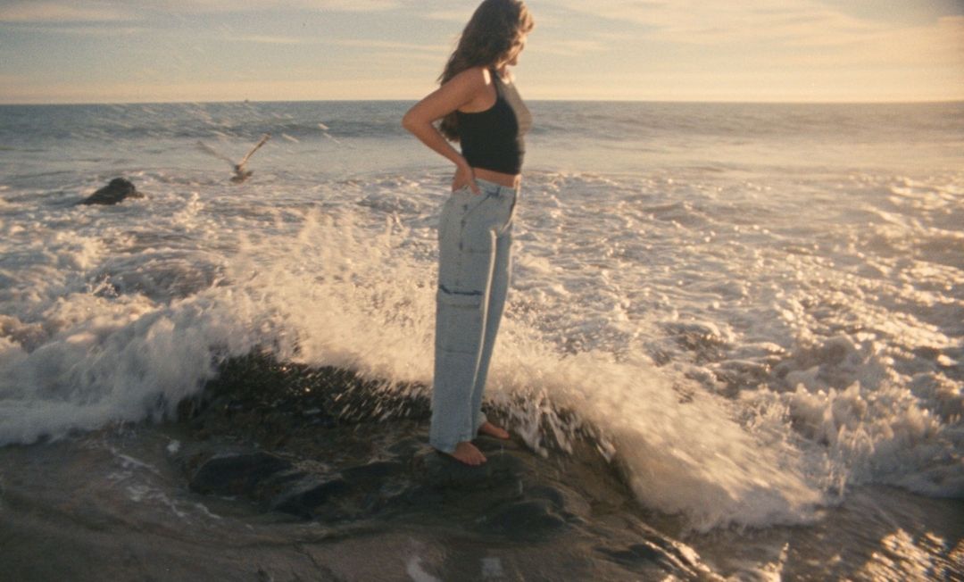 Woman in a black crop top and light wash jeans standing on a coastal rock as waves break around her at sunset.