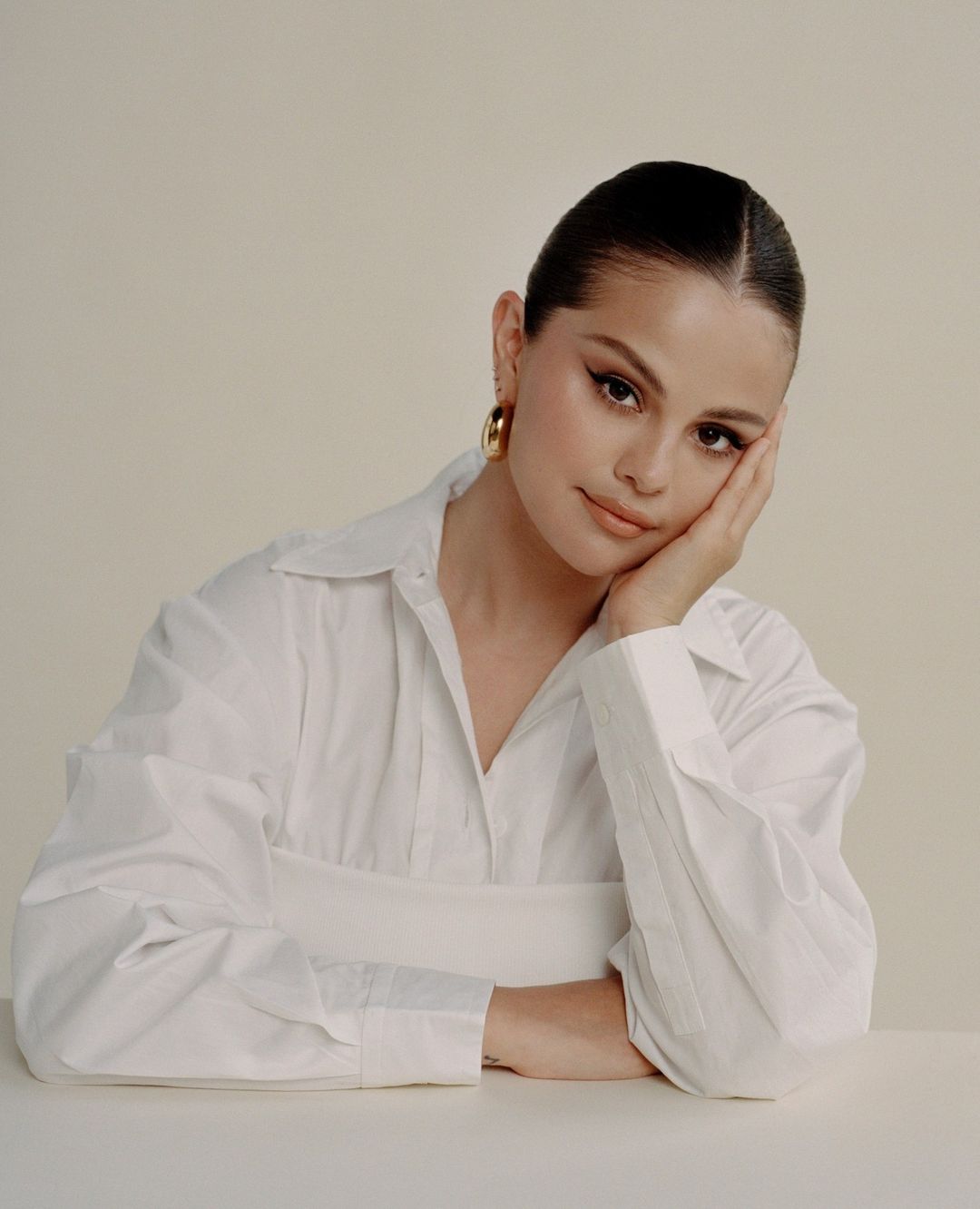 Portrait of Selena Gomez wearing a white button-down shirt and gold hoop earrings, resting her face on her hand, against a neutral background.