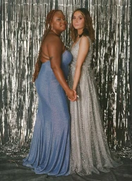 Two young women in formal evening gowns standing and holding hands against a shiny silver fringe backdrop in New York by Katie Mccurdy