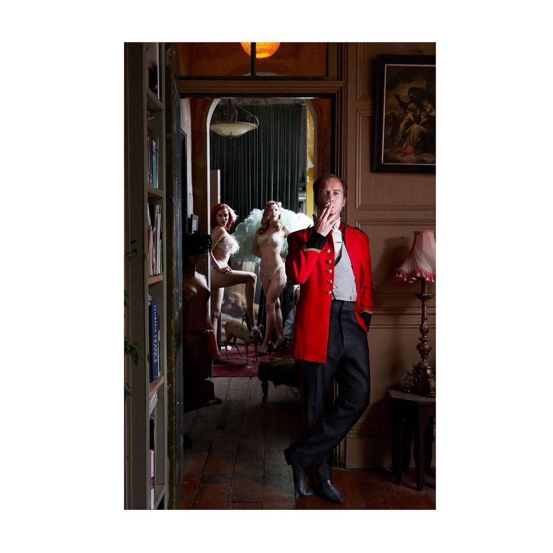 A man in a red jacket stands in a doorway, smoking a cigarette, while three women in lingerie pose in the background. Indoor setting.