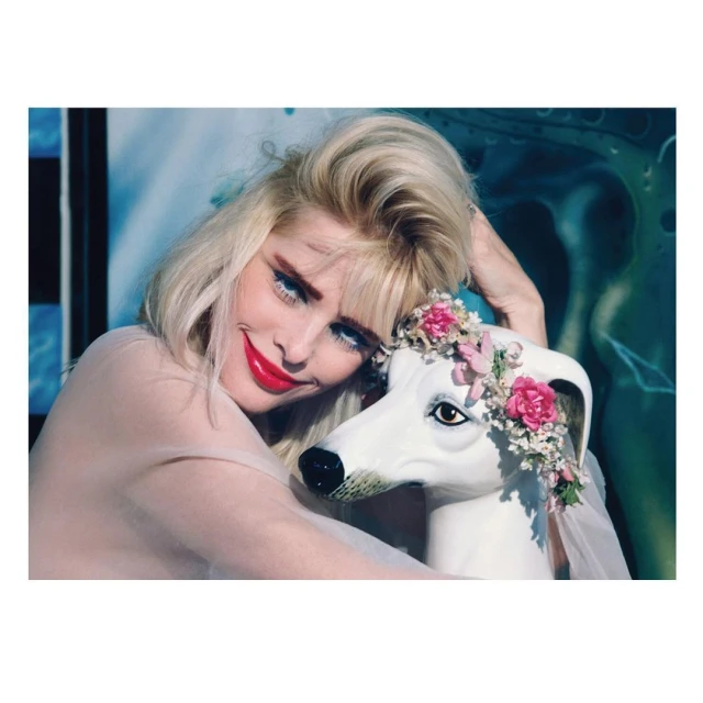 Woman with blonde hair smiles, embracing a decorative white dog bust adorned with flowers in a studio setting.
