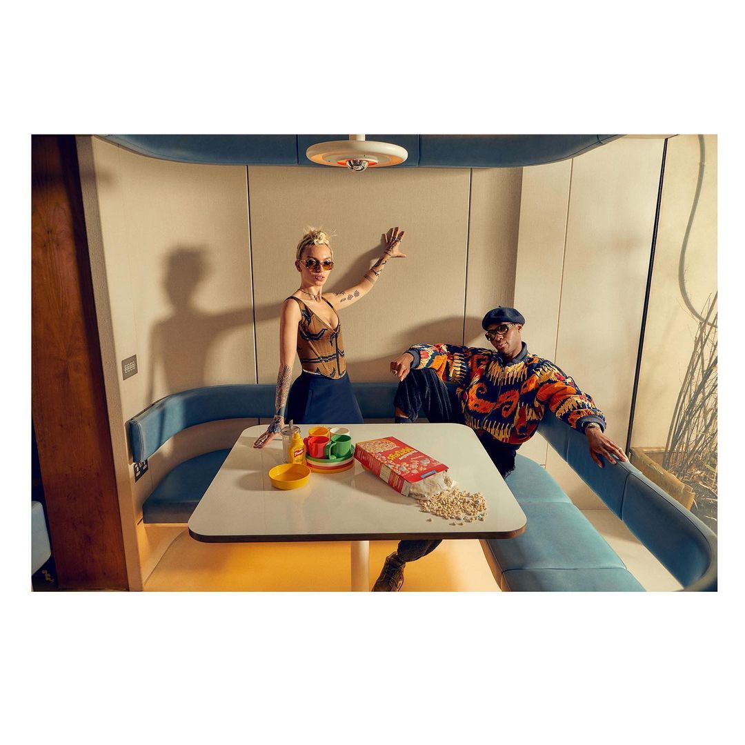 Indoor photo of a woman in corset and man in patterned sweater seated at a table with cereal box and scattered puffs.